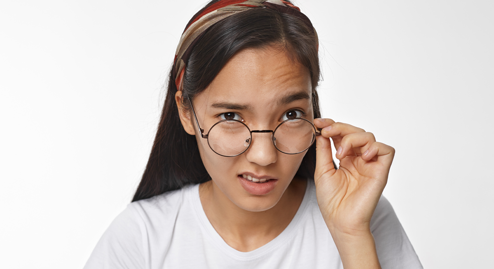 Portrait of a young woman with long dark hair, wearing a headband and white shirt, adjusting round glasses toward the camera in a studio setting.